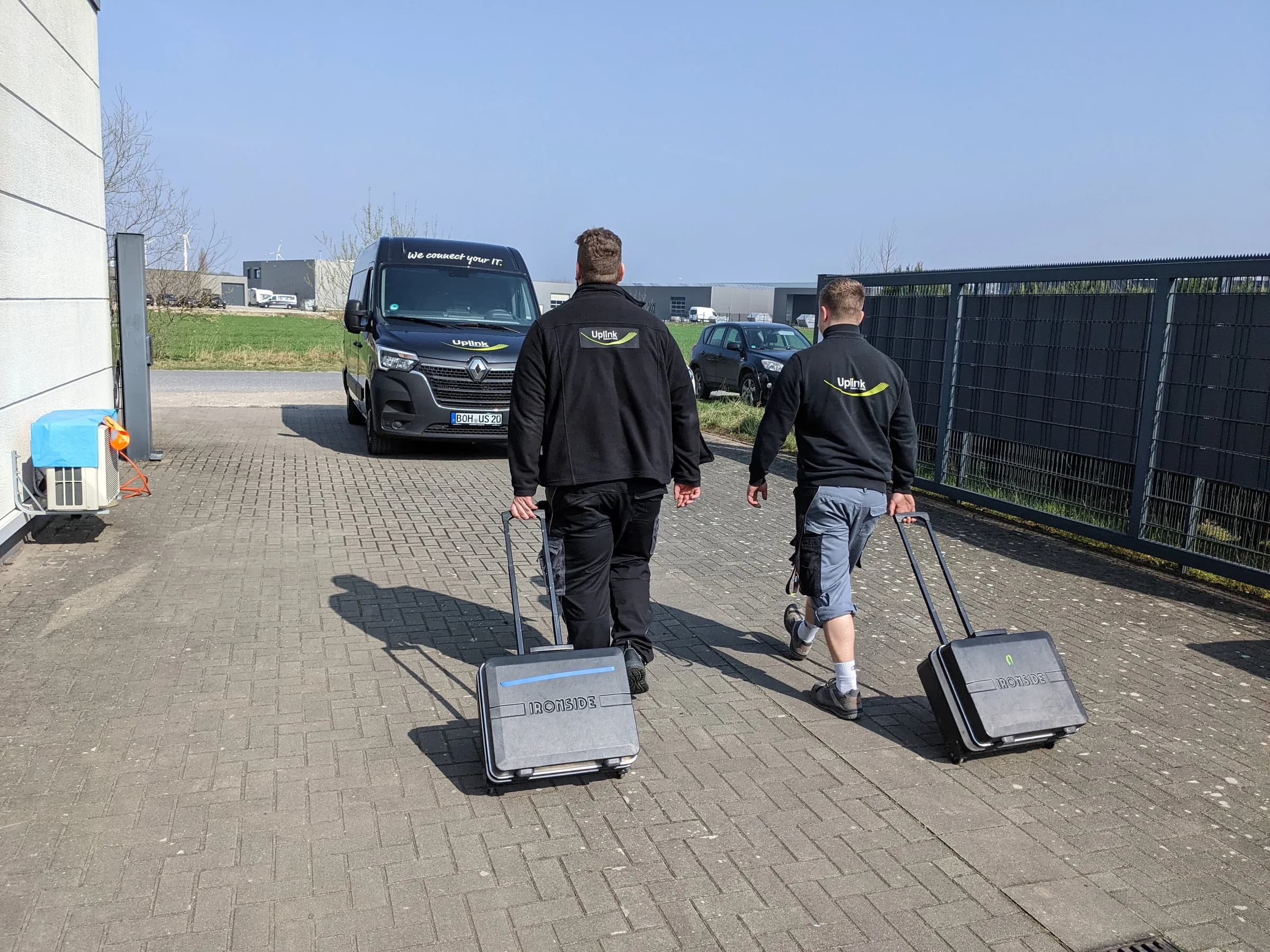 Zwei Uplink-Techniker ziehen Werkzeugkoffer über einen Hof zum Transporter