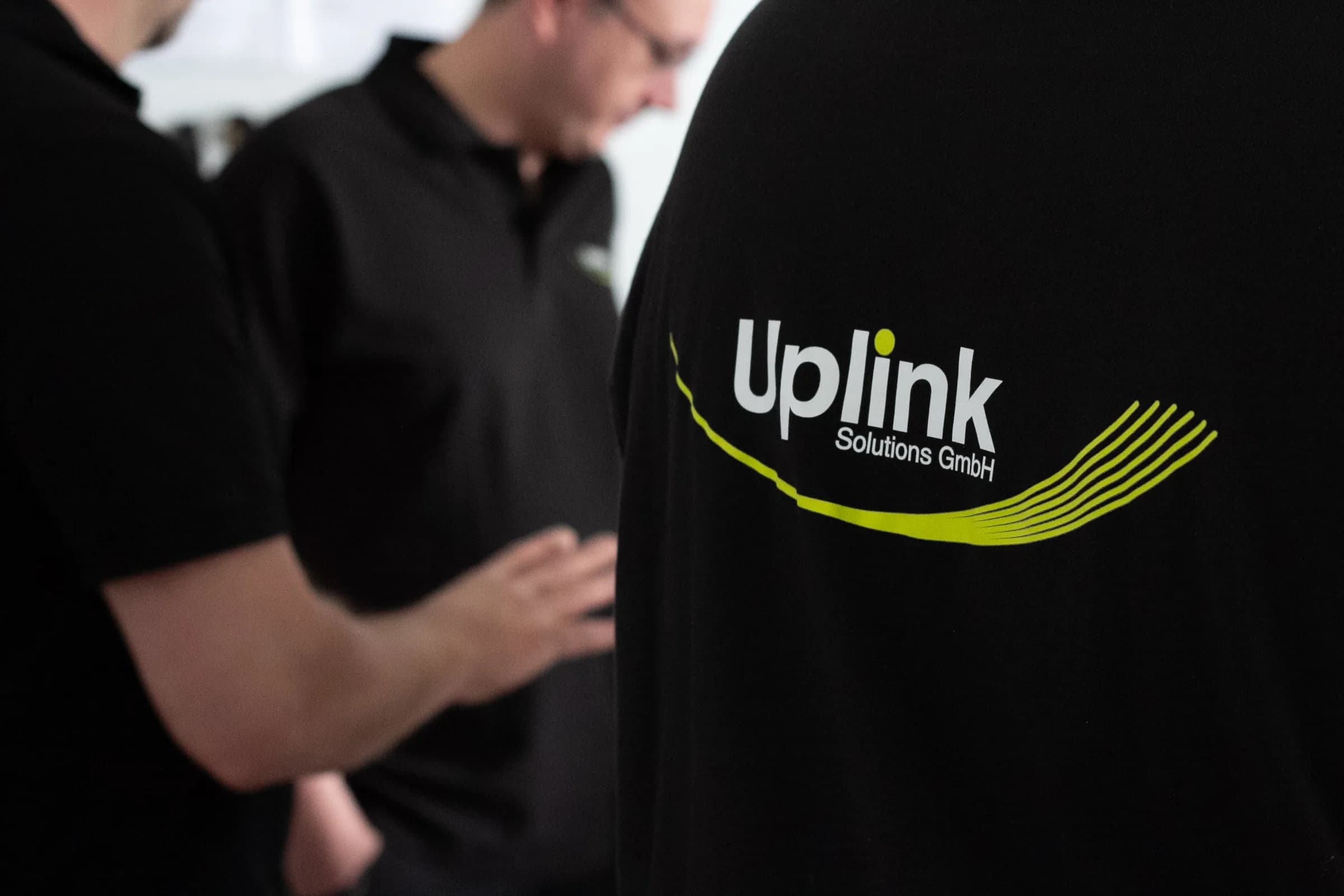 Nahaufnahme eines Technikers im schwarzen Poloshirt mit Uplink-Logo im Teamgespräch