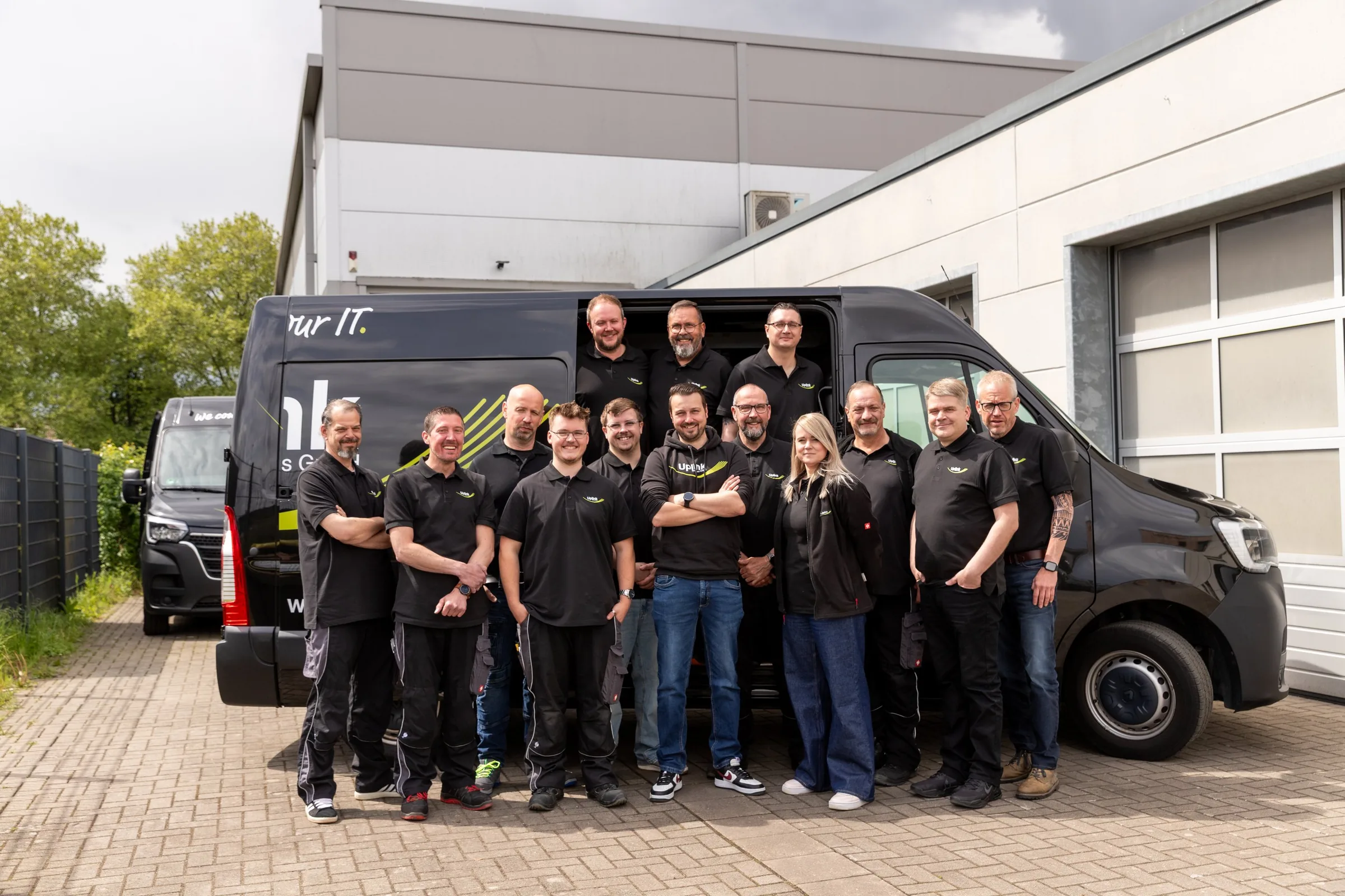 Das Uplink-Team in schwarzer Arbeitskleidung vor einem Firmenwagen auf dem Betriebsgelände